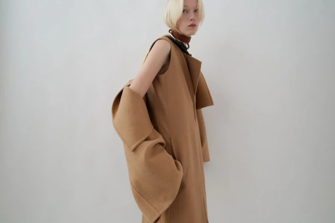 Fashion shot of a blonde woman in a tan, sleeveless coat, posing in front of a white background in a photo studio with soft lighting.