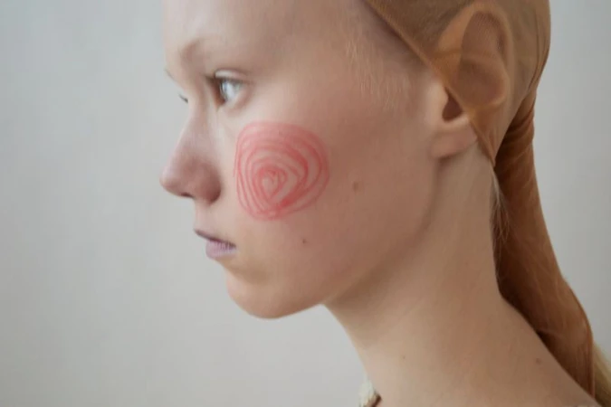 Profile of a person with fair skin and a blush heart-shaped makeup design on their cheek, wearing a tan head covering in a studio.