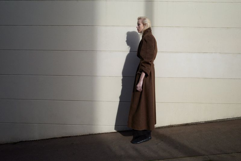 Model standing in profile wearing a long brown belted coat against a pale wall with a strong cast shadow