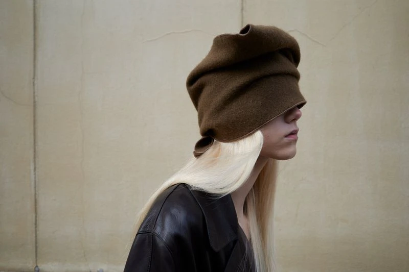 Profile portrait of a person wearing a brown head covering with long blonde hair and a black jacket, posing against a beige wall.