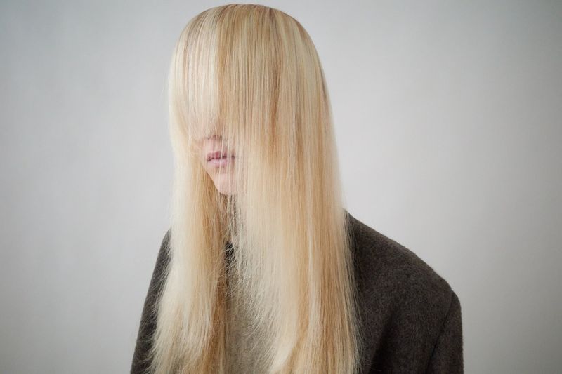 Close-up studio shot of a person with long blond hair covering their face, wearing a dark brown wool coat against a plain white background.