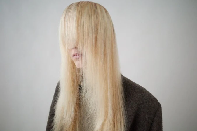 Close-up studio shot of a person with long blond hair covering their face, wearing a dark brown wool coat against a plain white background.