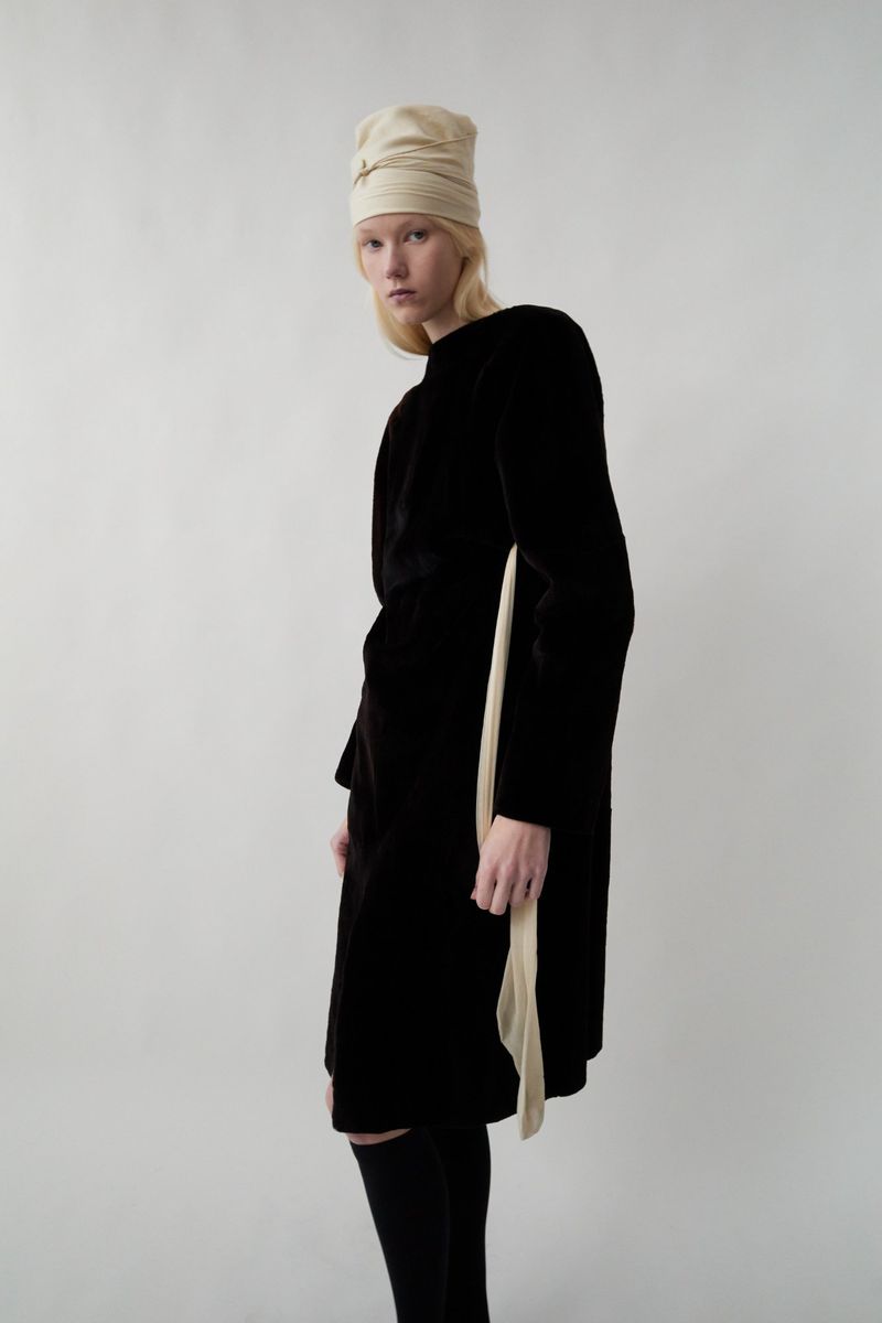 Young woman in black velvet dress and cream-colored head wrap posing in front of a plain white background in a fashion setting.