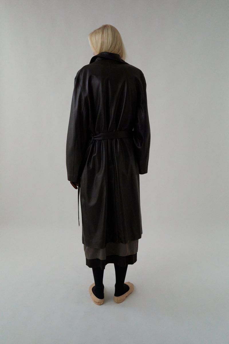 Model seen from behind wearing a dark leather belted coat against a plain white studio background.