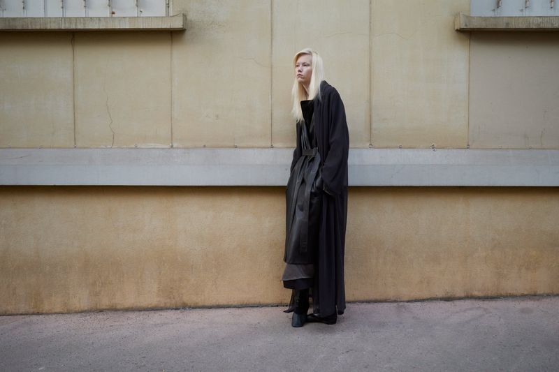 Blonde woman in dark coat stands against textured beige wall in an outdoor urban setting, fashionably dressed with long sleeves.