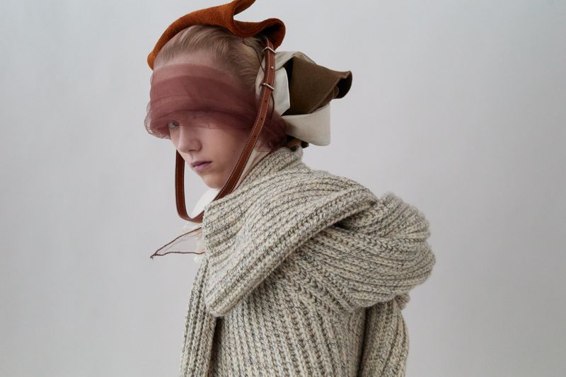Adult female in unique knitwear and abstract headwear poses in a studio. The neutral-toned fashion shot shows textured clothing elements.