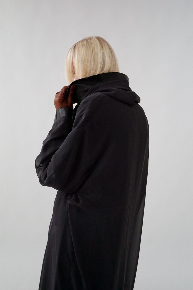 Back view of an adult female with blonde hair wearing a long, black coat with brown gloves against a plain grey studio backdrop.