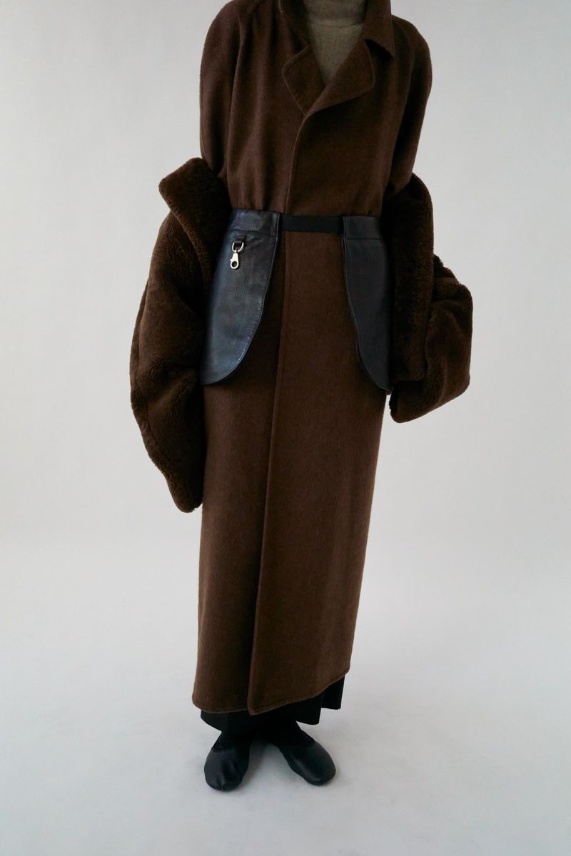 A person in a brown coat with fur sleeves and a black leather-like belt stands against a white background in a studio shot.
