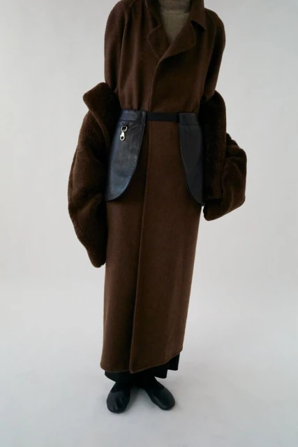 A person in a brown coat with fur sleeves and a black leather-like belt stands against a white background in a studio shot.