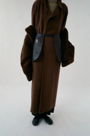 A person in a brown coat with fur sleeves and a black leather-like belt stands against a white background in a studio shot.