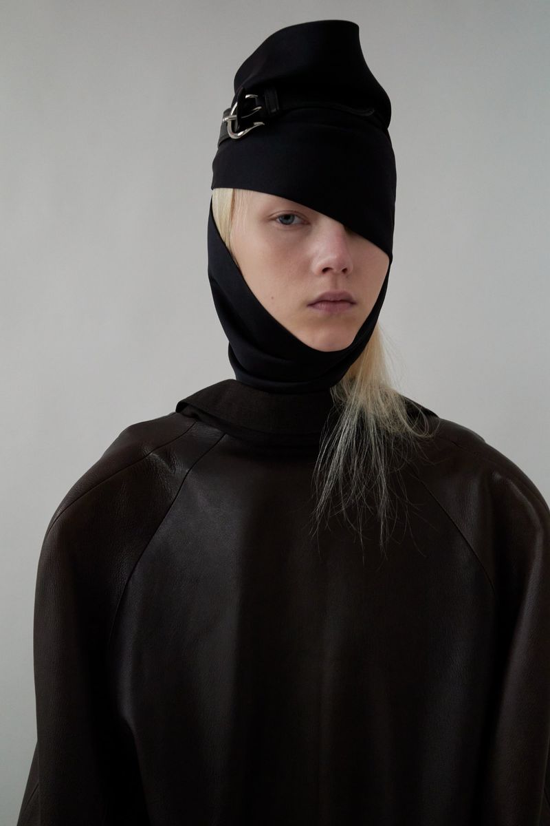 A solitary fashion portrait showcases a woman with blonde hair, styled with a black buckled head covering and a dark top against a pale background.