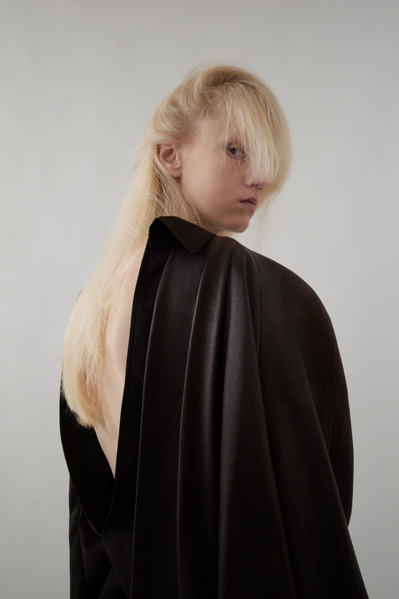 A solitary fair-skinned person with blonde hair, wearing a dark fashion top, gazes backward against a plain, light grey backdrop indoors.