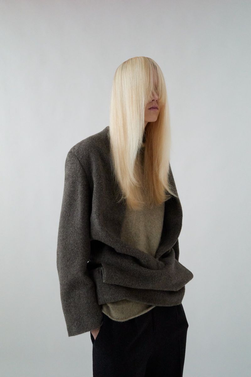 Fashion shot featuring an adult female model with straight blonde hair partially covering her face, posing in a studio with a gray coat.