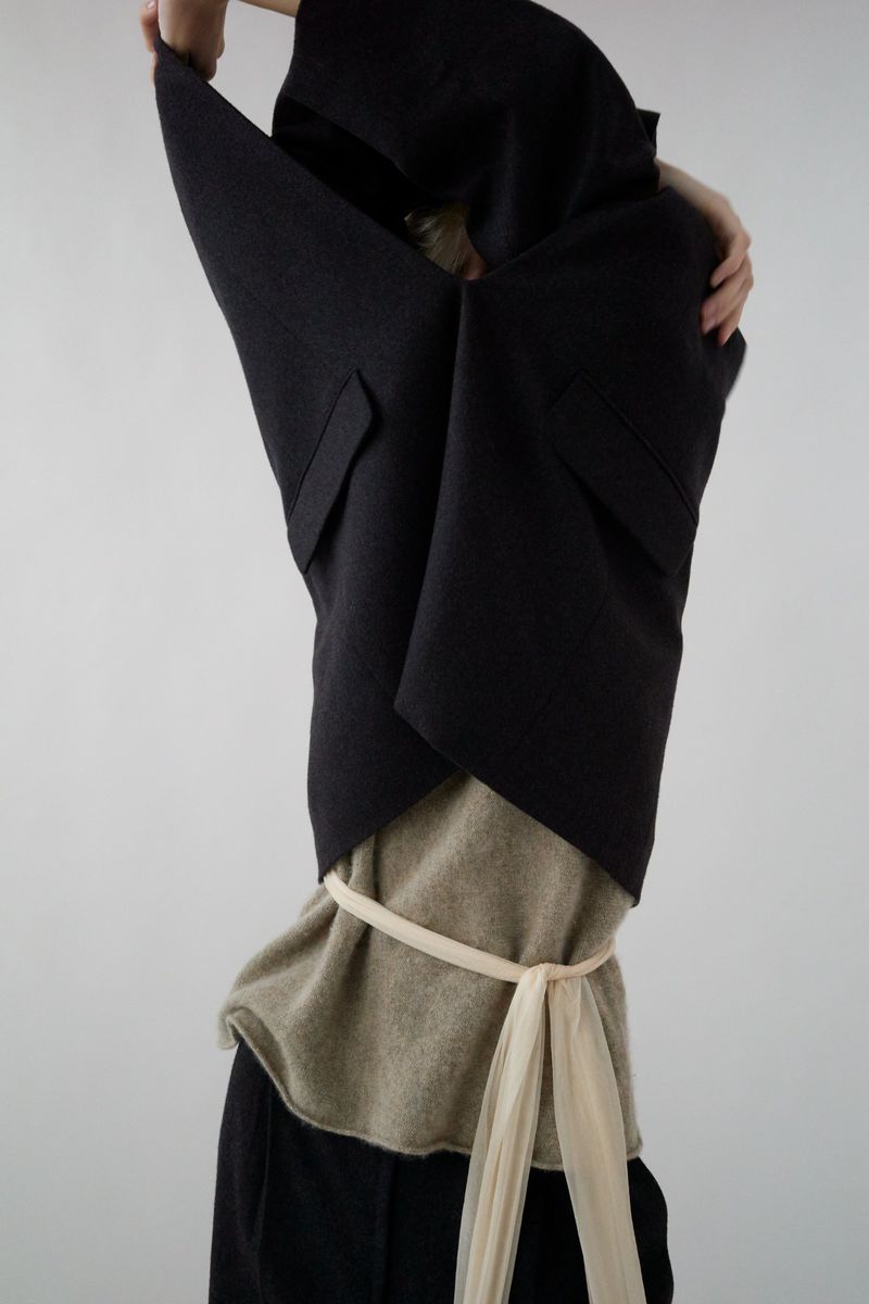 Fashion shot of a woman in a dark vest and neutral top, cinched with a light fabric belt, hands raised, against a bright neutral background.