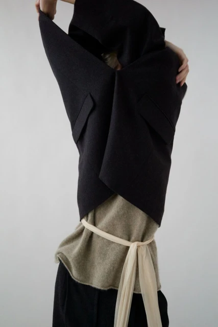 Fashion shot of a woman in a dark vest and neutral top, cinched with a light fabric belt, hands raised, against a bright neutral background.