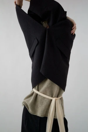 Fashion shot of a woman in a dark vest and neutral top, cinched with a light fabric belt, hands raised, against a bright neutral background.