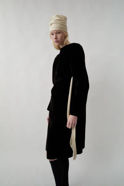 Model wearing a dark velvet dress and light beige draped headwrap against a plain white background