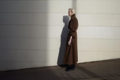 Model standing in profile wearing a long brown belted coat against a pale wall with a strong cast shadow