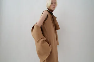 Model with blonde bob wearing a sleeveless camel wool coat against a plain white background