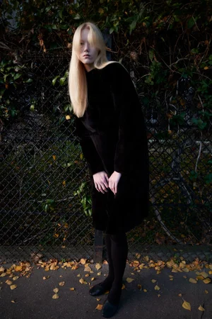 Model with long blonde hair wearing a black dress standing outdoors against a chain-link fence with foliage.