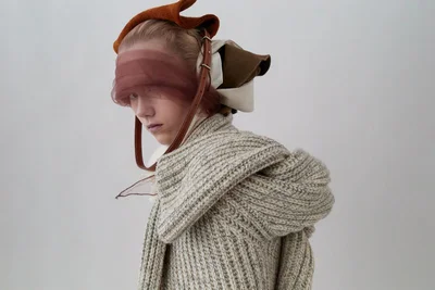 Model wearing a thick knit sweater and sculptural headwear wrapped in sheer fabric against a plain white background.