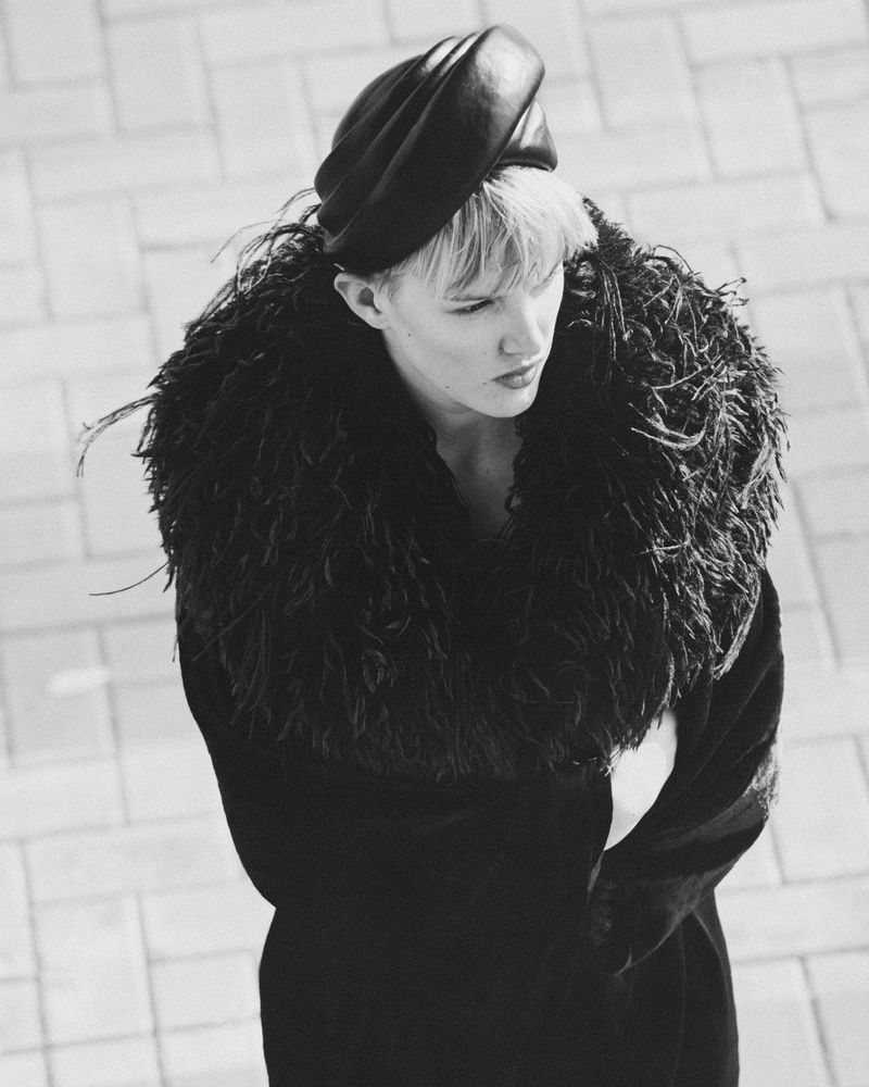 Black and white portrait of a stylish short-haired adult female wearing a beret and a feathered collar standing outdoors on a paved surface.