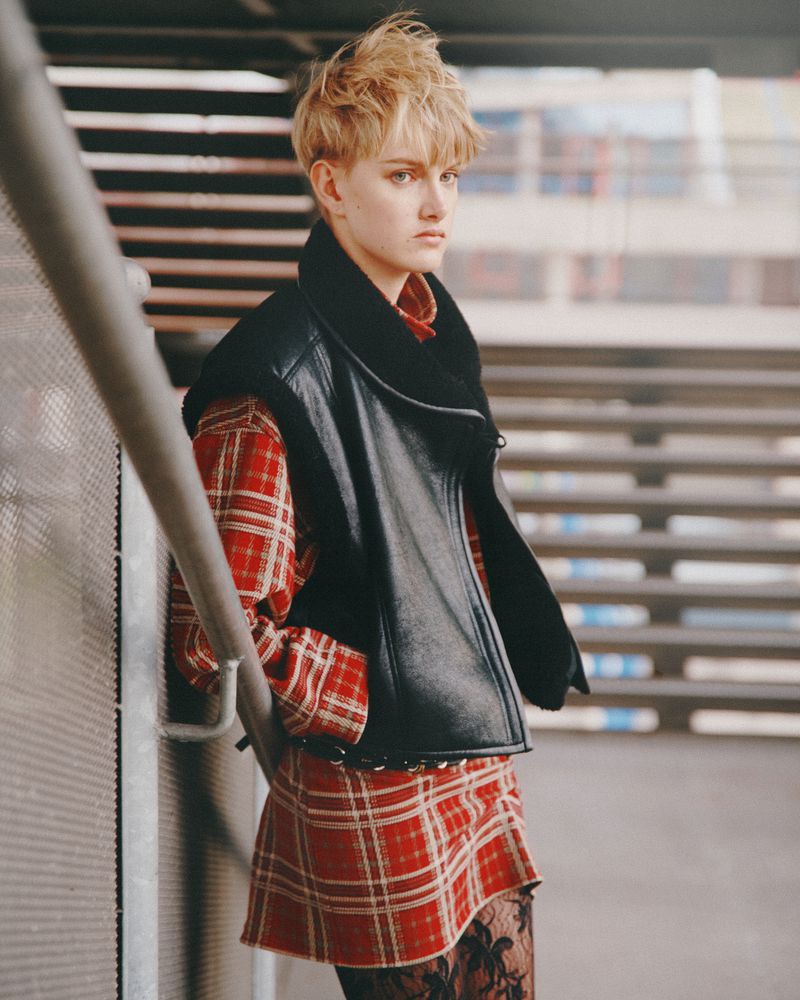 A fashionable young adult woman with short blonde hair poses on stairs wearing a black leather vest, red plaid dress, and black floral tights.