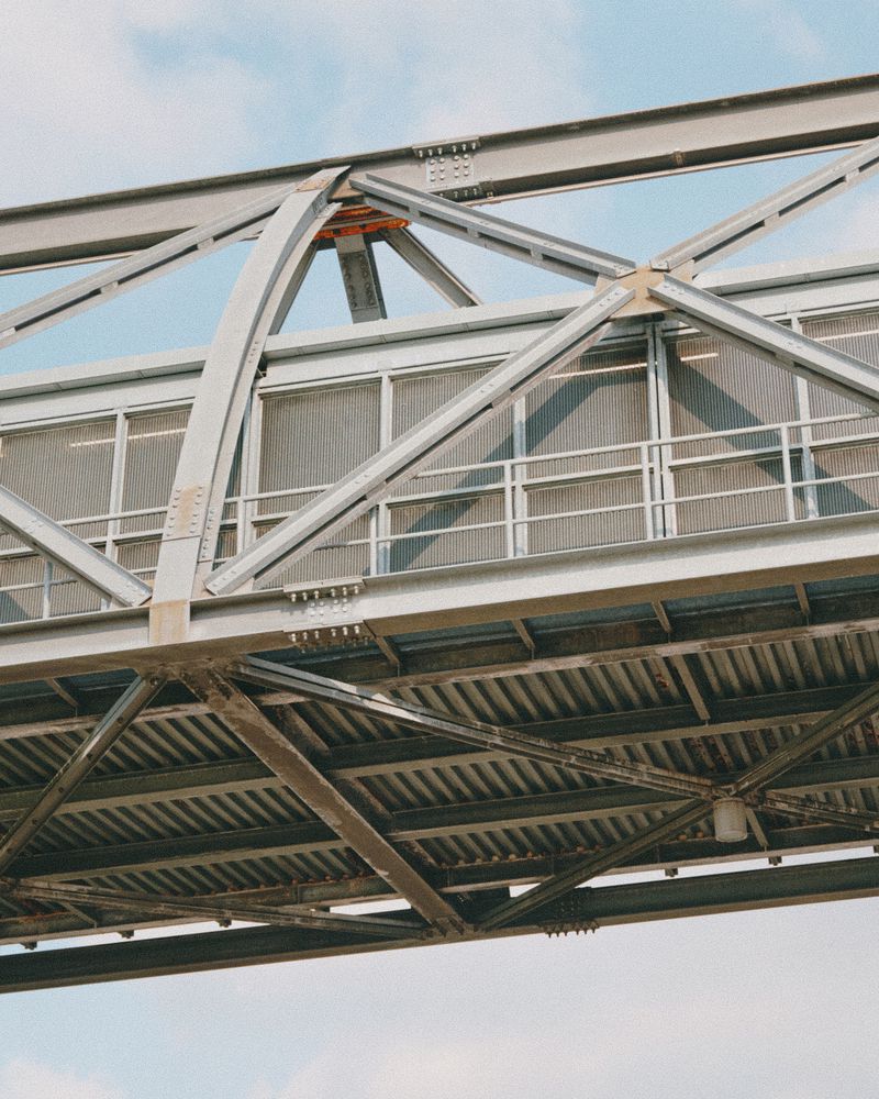 Low angle view of a gray steel overpass with lattice and beams against a blue sky on a sunny day, showcasing structural architecture.