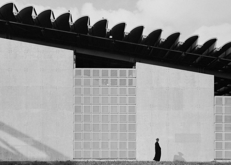 Architectural black and white photo features a lone woman walking beside a modern building with glass block windows under a cloudy sky.