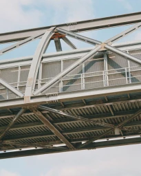 Low-angle upward view of an industrial steel bridge structure with crossbeams and railings against a pale blue sky.
