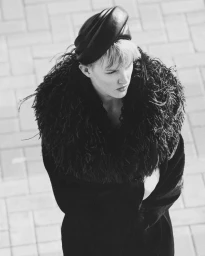 Model wearing a feathered collar coat and sculpted cap viewed from above against a paved brick ground.