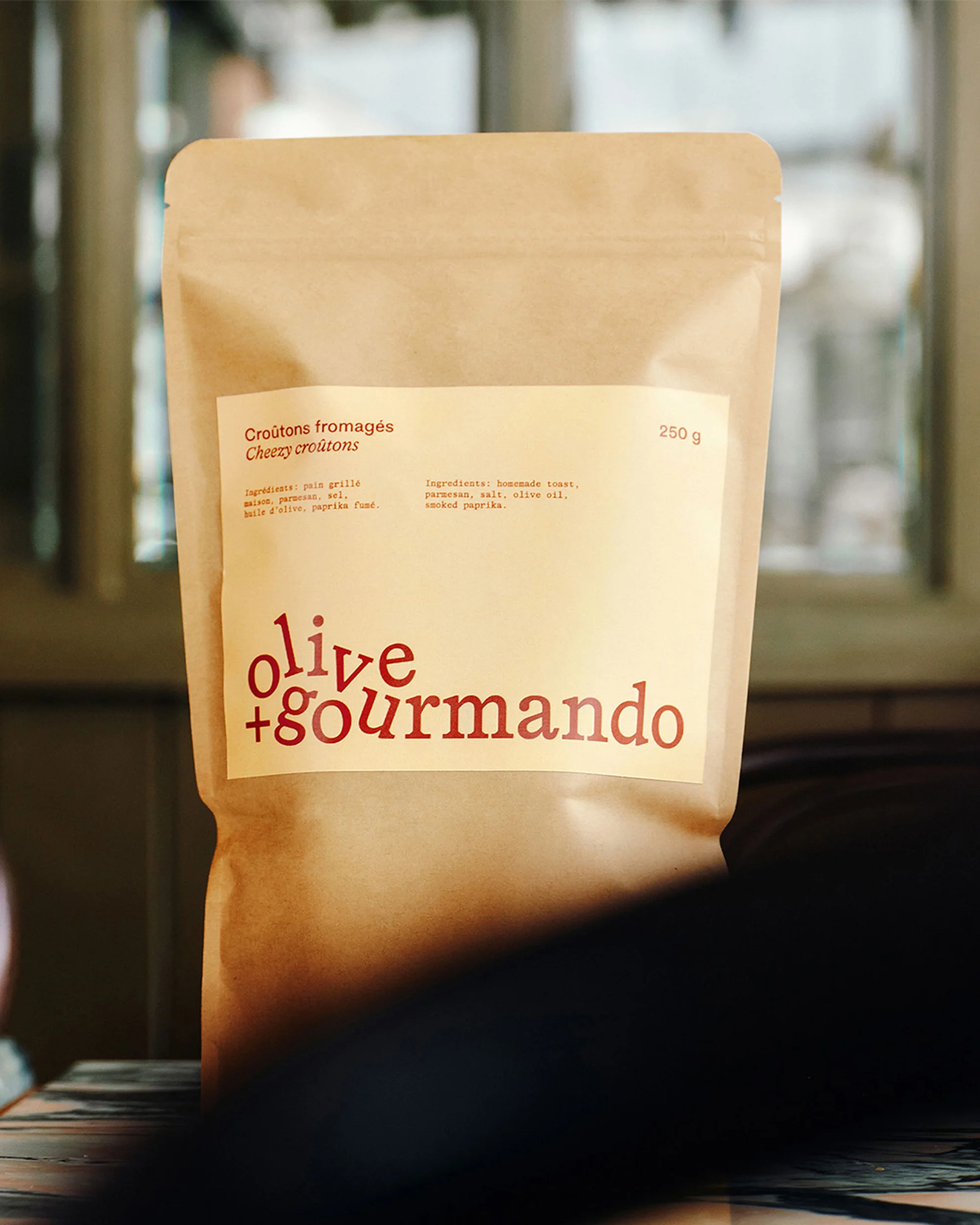 Olive + Gourmando cheese croutons in a paper bag on a table against a blurred window, promoting a savory food product.