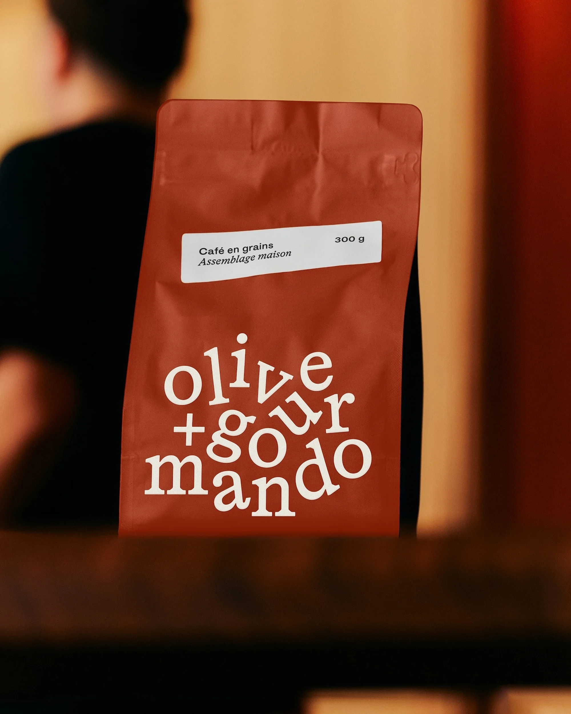 Eye-level shot of a brown coffee bag labeled 'Olive + Gourmando' sitting on a wooden surface, with a blurred figure in the background.