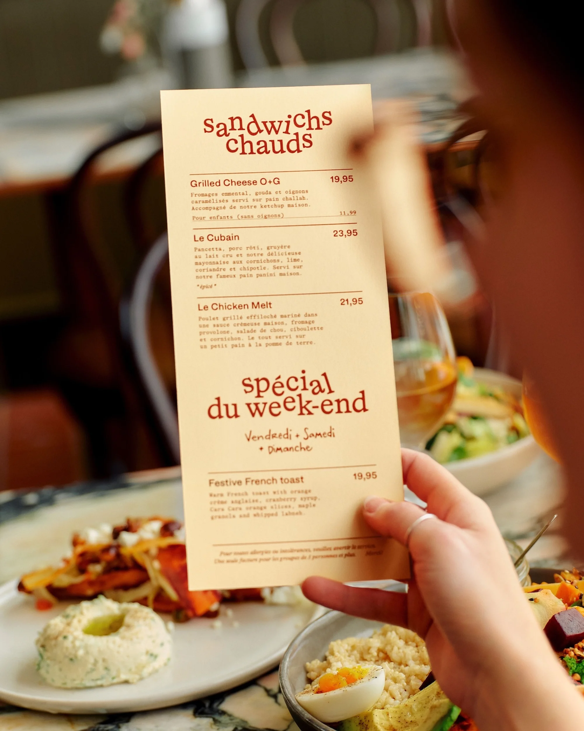 Person reads a menu with sandwich and French toast options while seated at a restaurant table with plates of food.