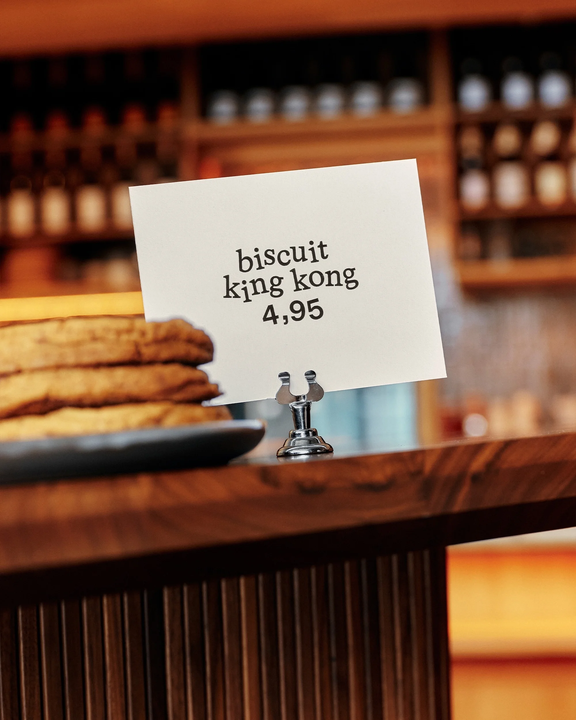 Close-up of cookies and a sign on a wooden counter in a cafe. The sign reads 'Biscuit King Kong 4,95'. Warm, inviting atmosphere.