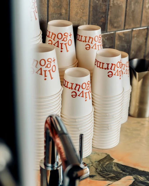 Stacks of white paper cups with 'live + gourmet' print, arranged neatly on a marble countertop against a tiled wall backdrop.