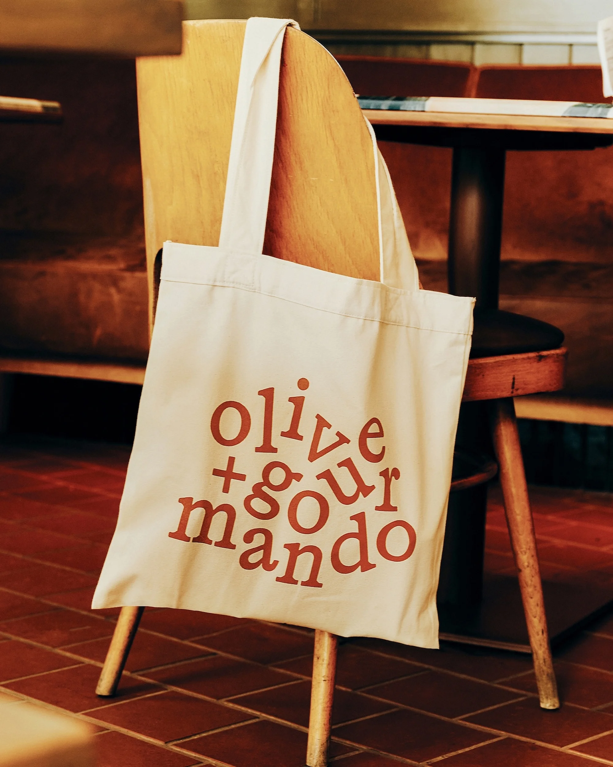 Tote bag with 'olive + gourmando' text leaning on a wooden chair inside a cafe, with wooden table and floors in the background.