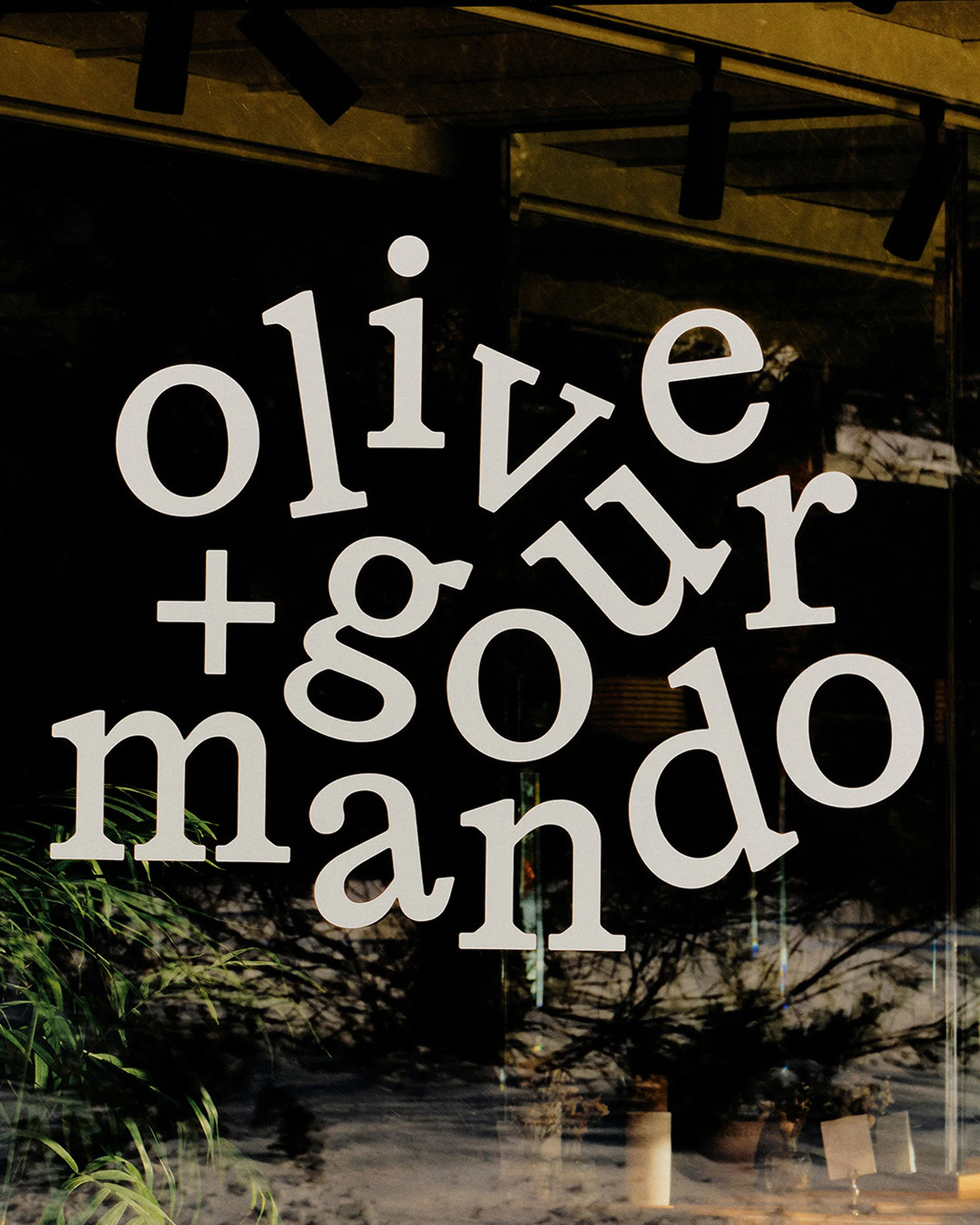 Olive + Gourmando signage on a window with greenery in the background. The text is in a sans-serif font and white color.