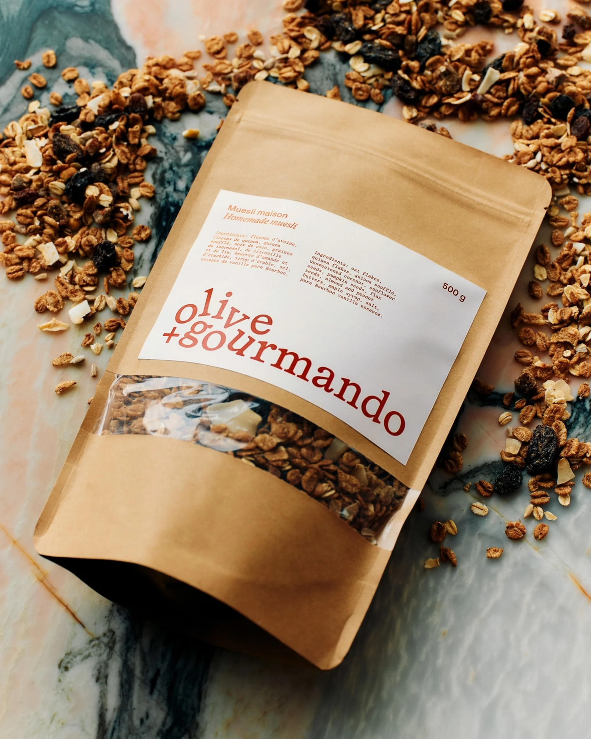 Granola in a branded brown bag with a clear window, scattered on a marble surface, featuring a mix of oats, nuts, and dried fruit.