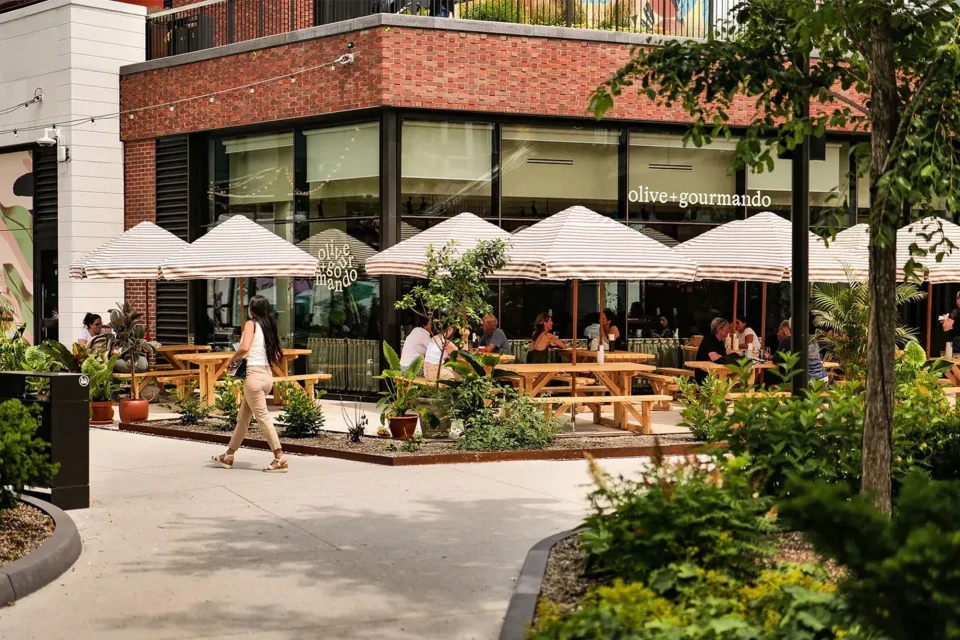 Outdoor cafe scene with patrons dining under umbrellas, featuring a woman walking by and lush greenery in an urban setting.
