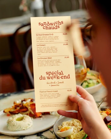Close-up of a hand holding a cream-colored restaurant menu with brown typography, set against a blurred dining background.