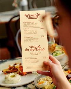 Close-up of a hand holding a cream-colored restaurant menu with brown typography, set against a blurred dining background.