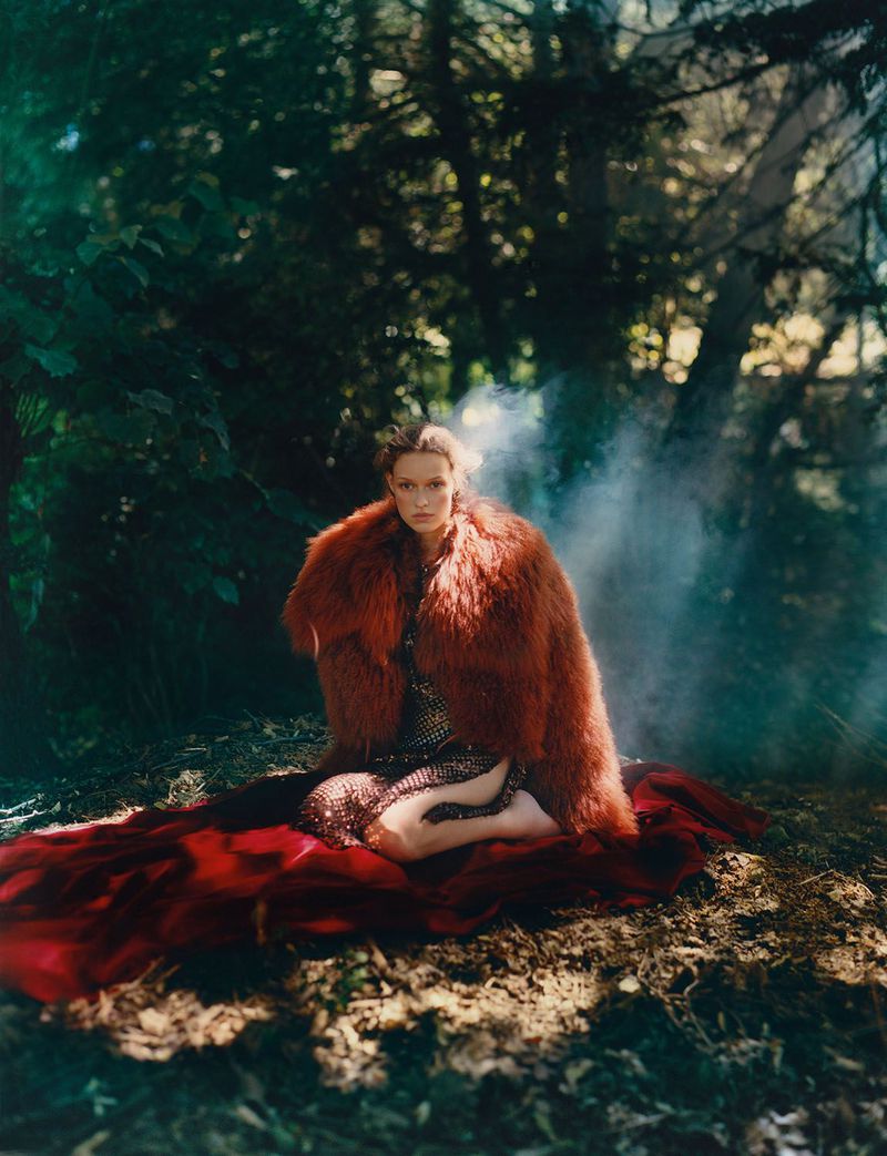 A woman in an orange fur coat sits on a red fabric in a forest with dappled sunlight and a hazy background, showcasing fashion and style.