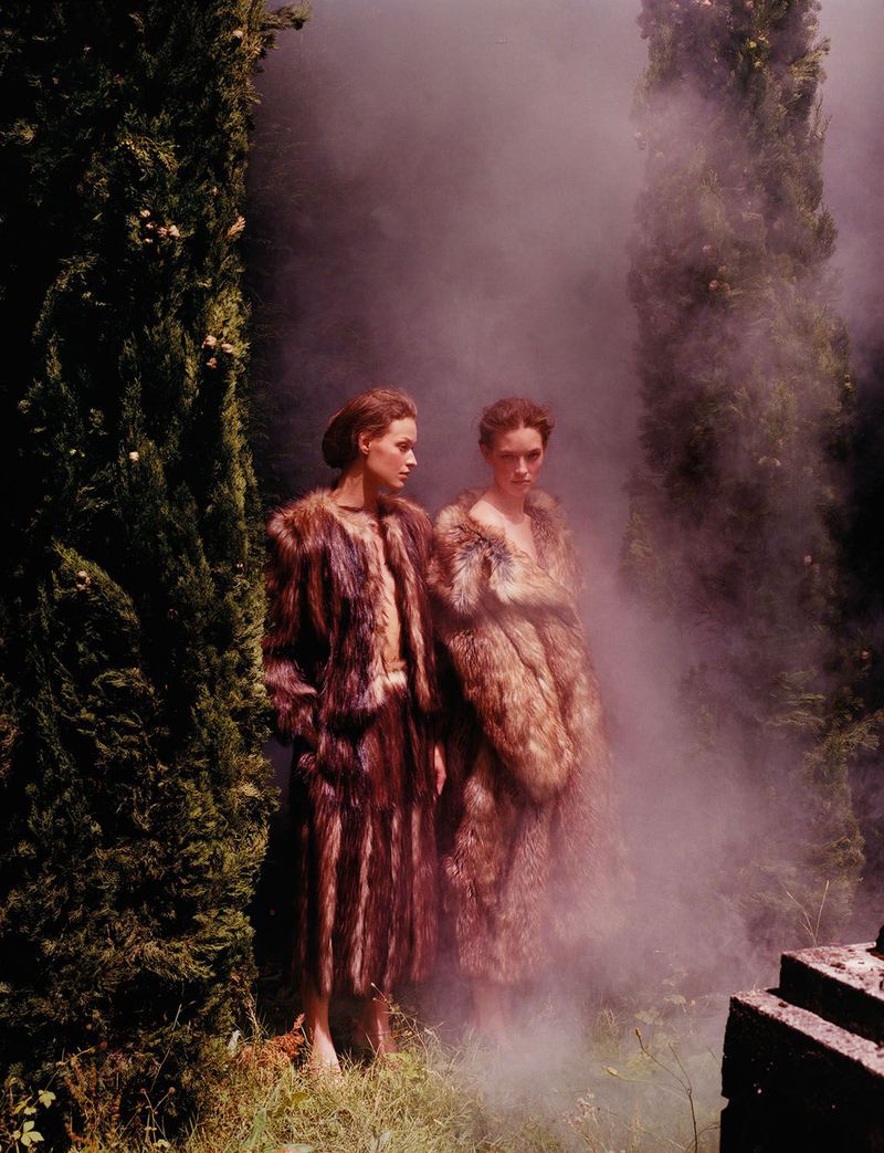 Two women in fur coats stand amidst an ethereal, smokey outdoor setting with tall trees, evoking a sense of high fashion and mystery.
