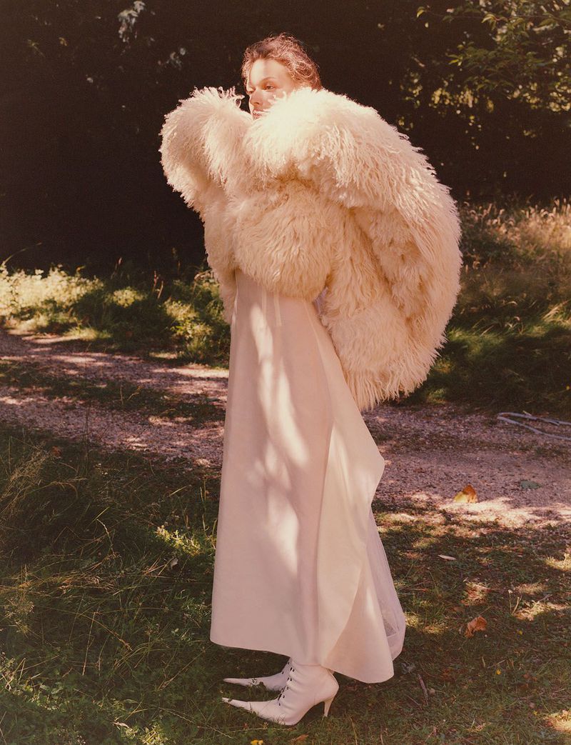A woman wearing a large fur coat and long dress stands in a grassy outdoor setting, showcasing a fashion-forward editorial look.
