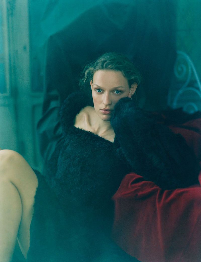 Portrait of a woman with curly hair wearing a dark fur coat indoors. A red blanket adds contrast. Soft, diffused lighting creates a moody atmosphere.