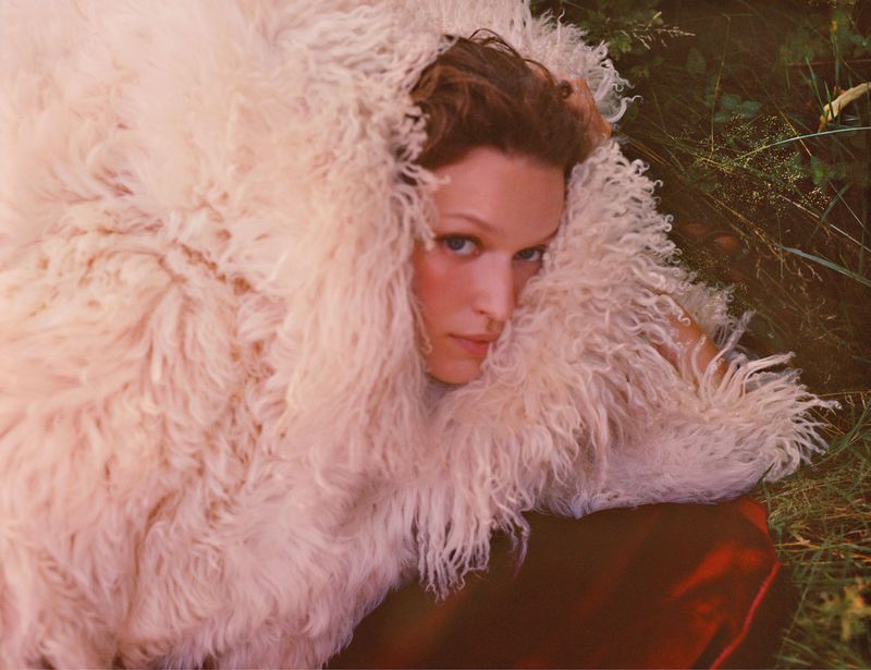 Portrait of a woman with fair skin, brown hair, and blue eyes wrapped in a fluffy, cream-colored garment against a natural backdrop.