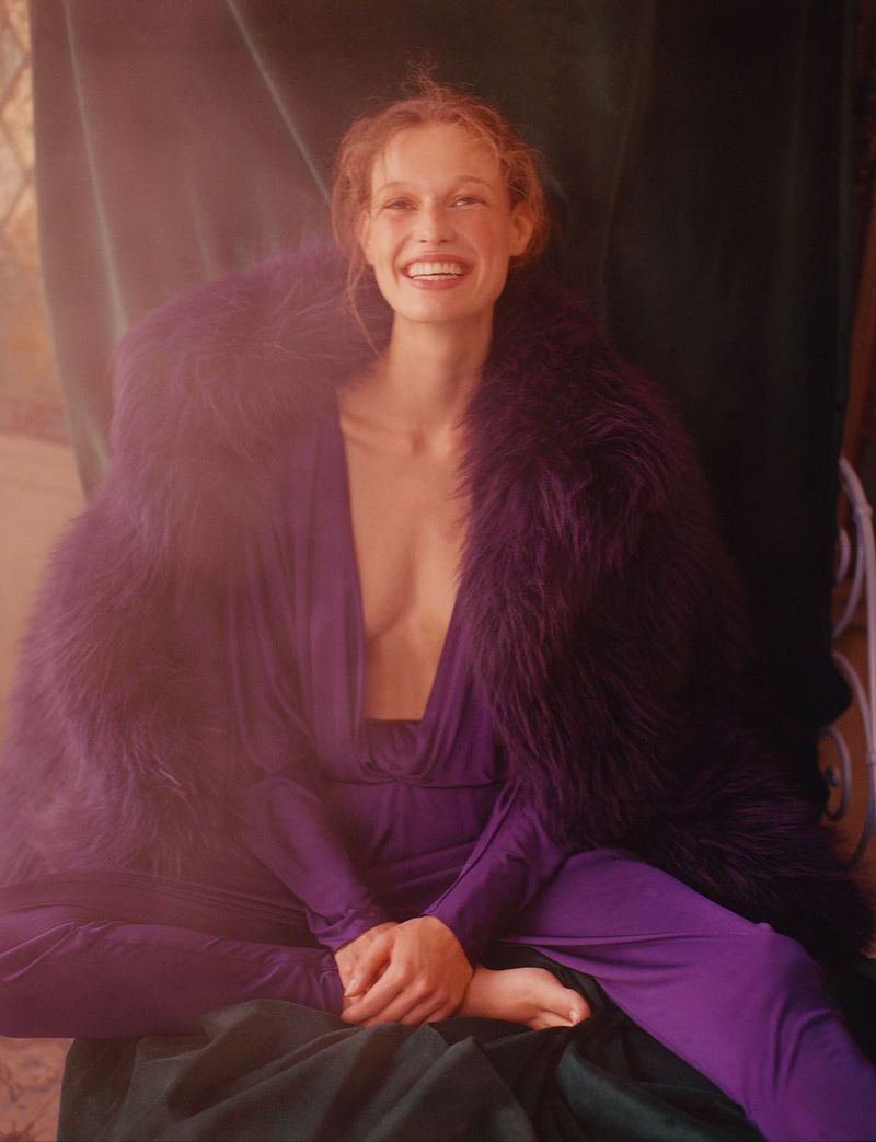 Woman in purple dress and faux fur shawl smiles indoors against a green draped backdrop with a window casting natural light.