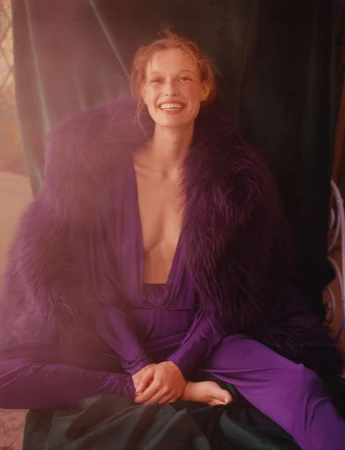 Woman in purple dress and faux fur shawl smiles indoors against a green draped backdrop with a window casting natural light.