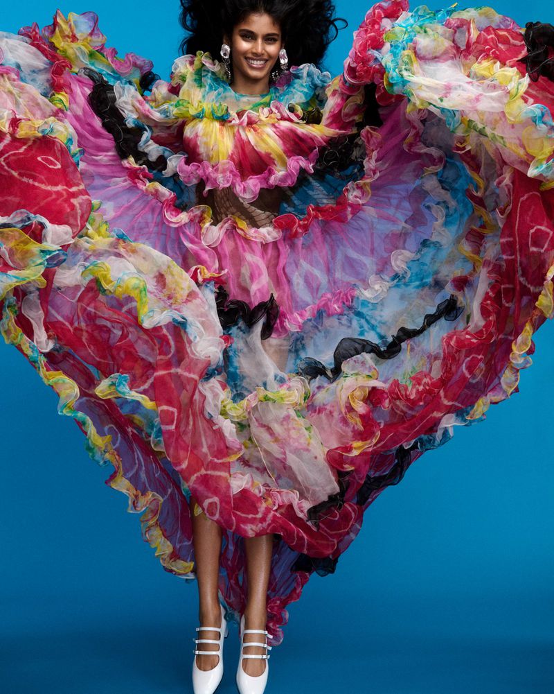 Model jumping with arms spread wide wearing a voluminous vibrant multi-colored ruffled dress against a solid blue backdrop